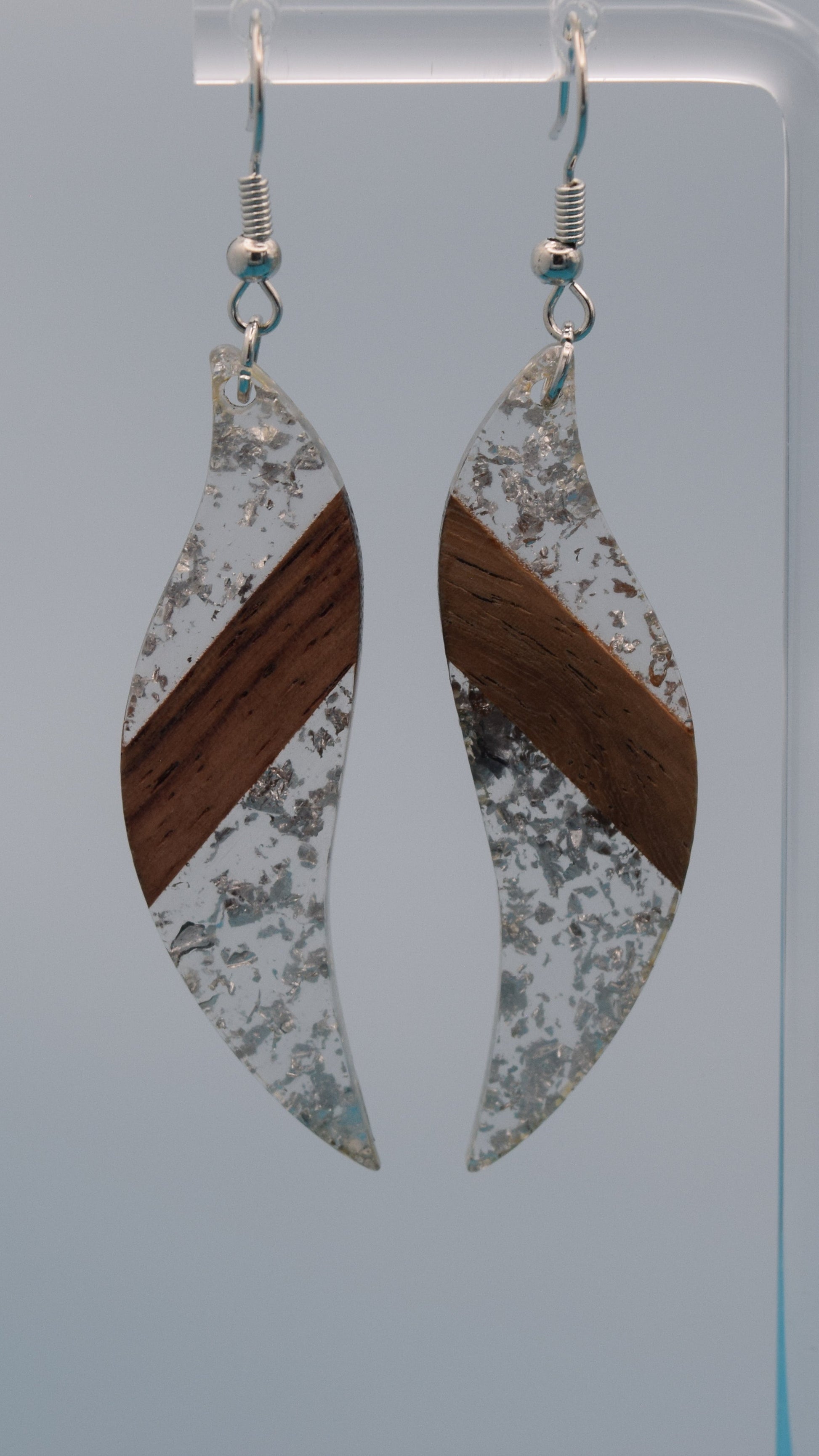 The earring is a translucent resin. This resin has a smooth, polished finish, allowing some light to pass through, giving it a luminous quality. Embedded within the resin are numerous small, irregular silver flakes with a walnut color piece of wood set in the middle of the earring. These flakes are scattered randomly, catching the light and adding a subtle, delicate shimmer and texture to the surface.