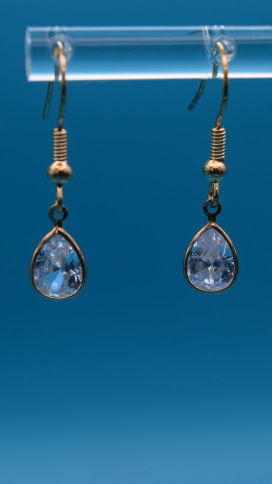 Teardrop-shaped dangling earrings hanging at 1/2 inch long featuring gold teardrop edge with a clear crystal incased at the center. Displayed on a clear stand against a teal background.