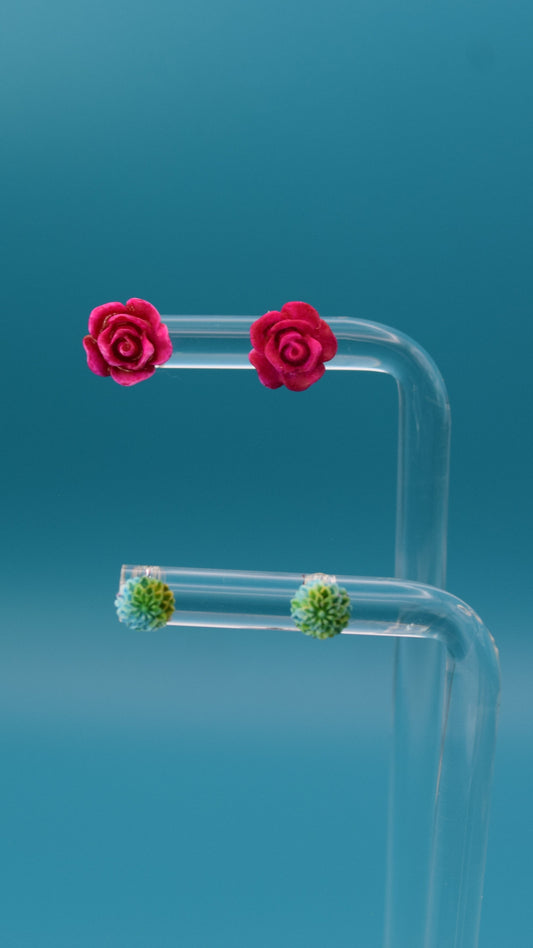 Hot pink rose-shaped stud earrings, paired with a smaller dahlia bloom featuring blue and green coloring. Displayed on a clear stand against a teal background.