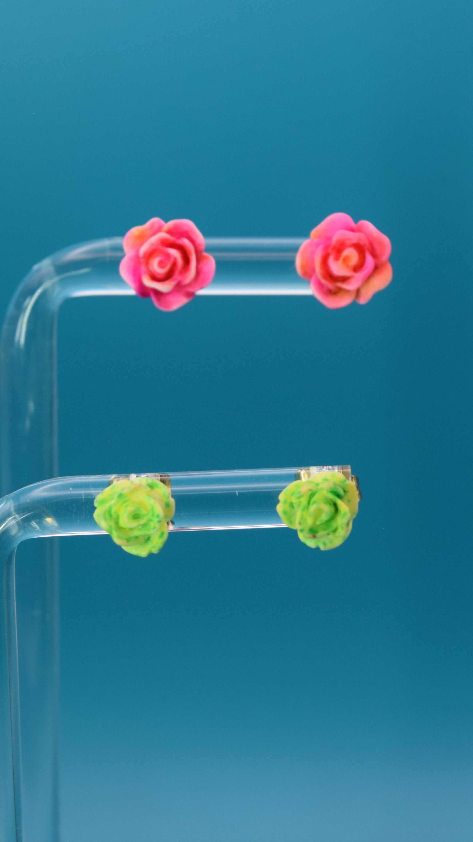 Hot pink and orange tie dyed effect rose-shaped stud earrings, paired with a smaller yellow and green tie dyed effect rose. Displayed on a clear stand against a teal background.