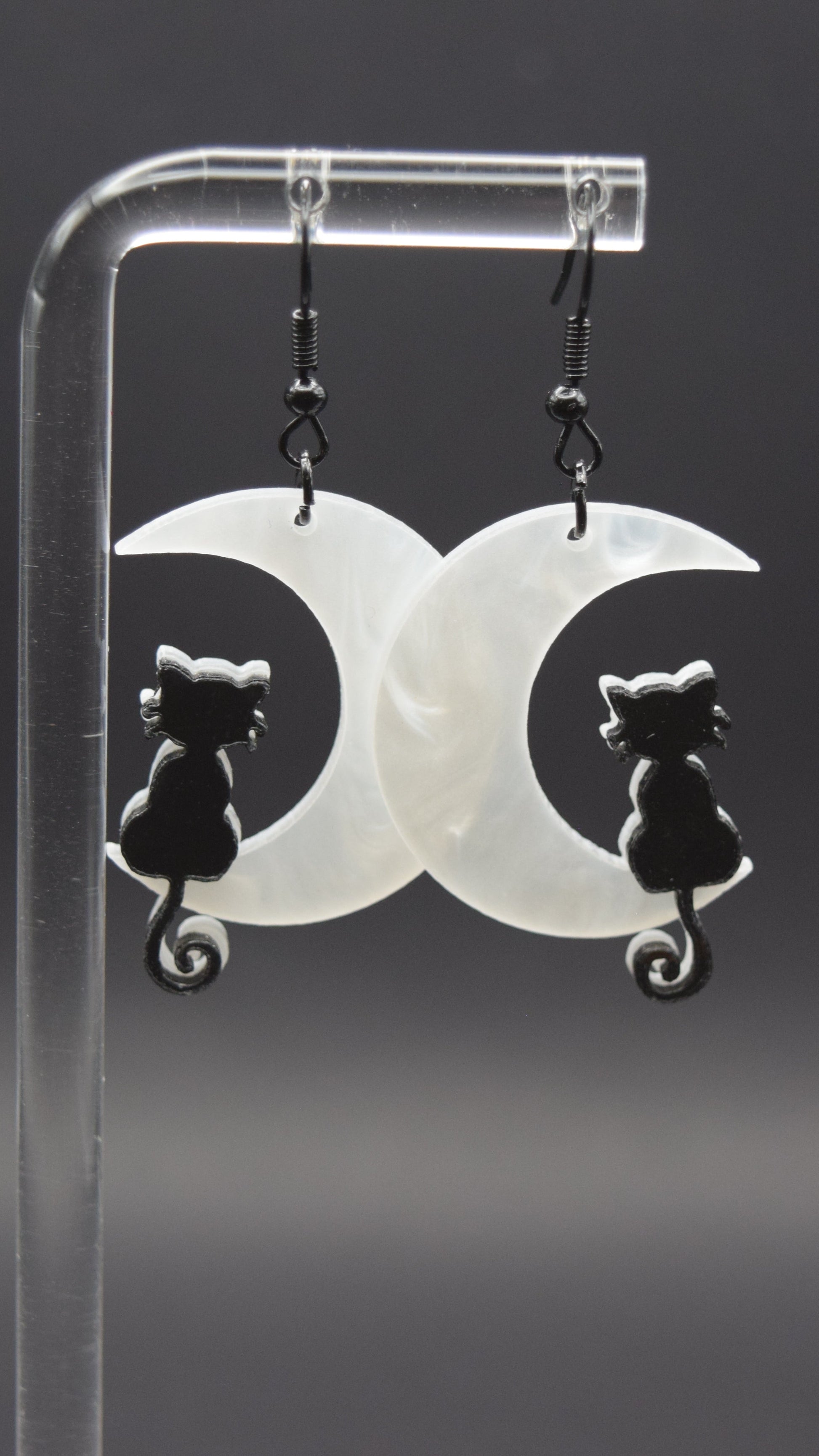 Each earring consists of a white, opalescent acrylic crescent moon facing to the left, with a smooth, slightly wavy texture visible in the material. Sitting on the lower curve of each moon is a black, silhouetted cat. The cats are facing away from the viewer, with their tails curled into a spiral at the base. Displayed from a clear, acrylic stand against a dark background.
