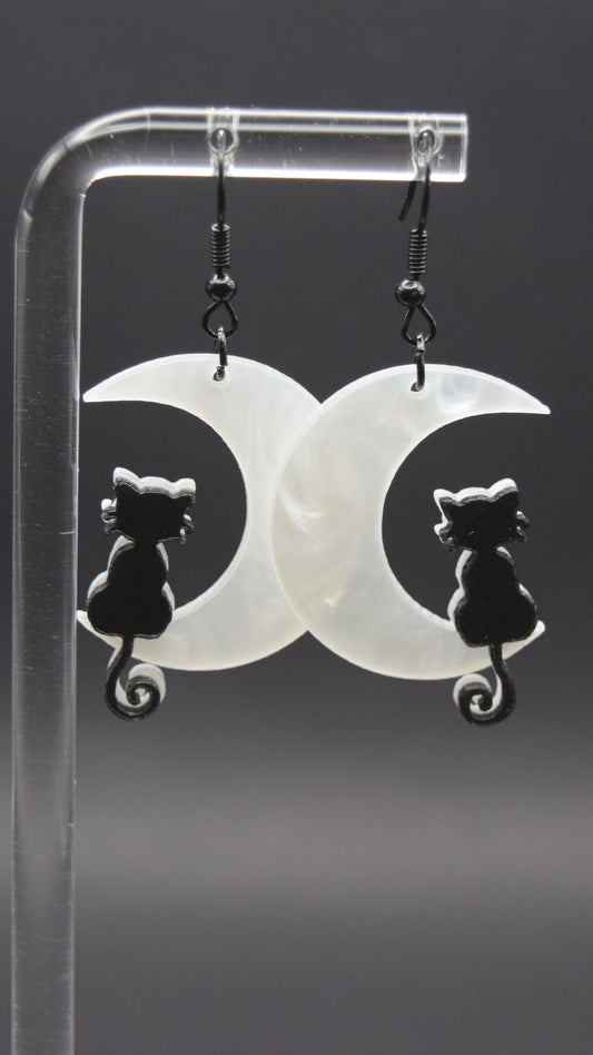 Each earring consists of a white, opalescent acrylic crescent moon facing to the left, with a smooth, slightly wavy texture visible in the material. Sitting on the lower curve of each moon is a black, silhouetted cat. The cats are facing away from the viewer, with their tails curled into a spiral at the base. Displayed from a clear, acrylic stand against a dark background.