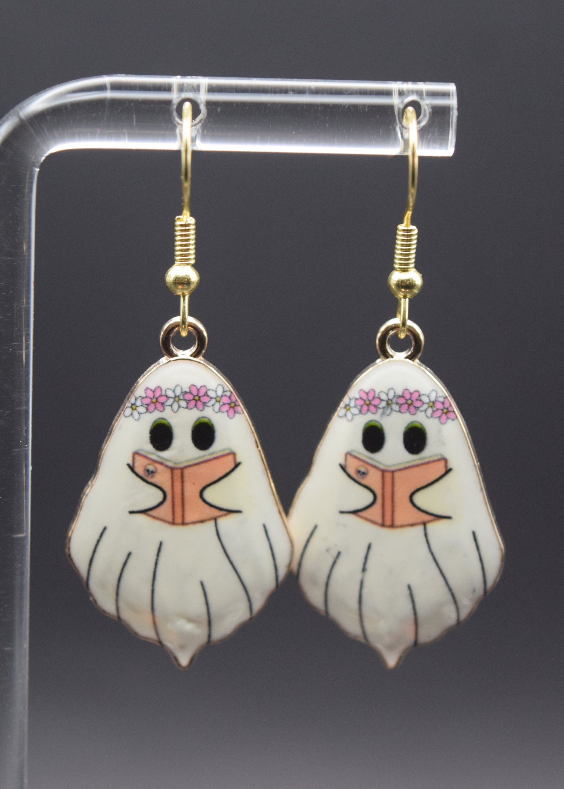 Each earring features a ghost-like figure with a slightly whimsical and friendly appearance. The main body of each ghost is white with faint vertical lines to suggest movement or drapery. The ghosts have black outlines. At the top of each ghost's head, there's a delicate floral crown, predominantly pink and white. Below the crown, each ghost has two small black eyes. Instead of a typical mouth, there's an open book design, with pale orange pages and black lines making it appear as if the  ghost is reading.