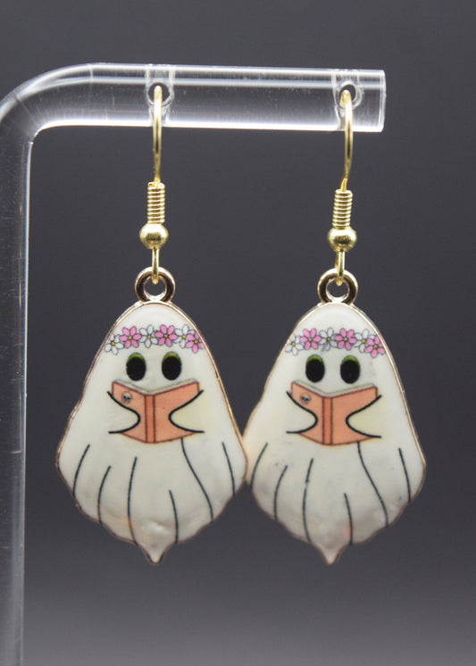 Each earring features a ghost-like figure with a slightly whimsical and friendly appearance. The main body of each ghost is white with faint vertical lines to suggest movement or drapery. The ghosts have black outlines. At the top of each ghost's head, there's a delicate floral crown, predominantly pink and white. Below the crown, each ghost has two small black eyes. Instead of a typical mouth, there's an open book design, with pale orange pages and black lines making it appear as if the  ghost is reading.