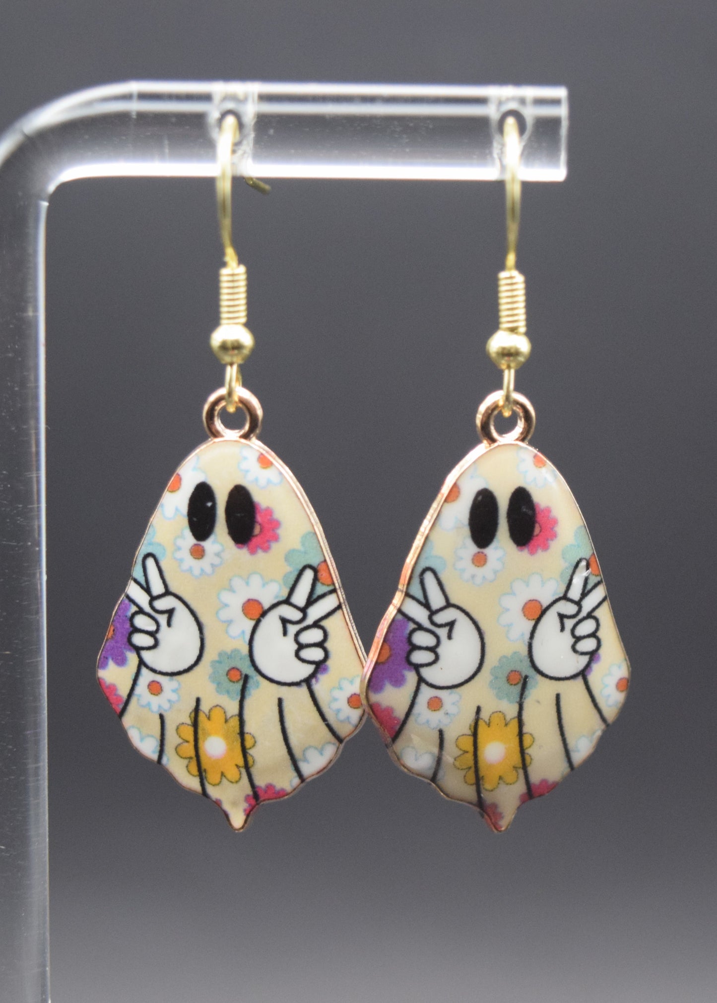 Each earring is shaped like a ghost, with a off-white base color. The entire surface of the ghost is adorned with a pattern of small, flowers in shades of yellow, white, blue, pink, and purple,. Each ghost has two black, oval-shaped eyes. In the center of each ghost, two white cartoon hands emerge, each making a "peace sign" gesture with the index and middle fingers raised in a 'V' shape. The hands have black outlines.