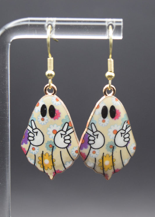 Each earring is shaped like a ghost, with a off-white base color. The entire surface of the ghost is adorned with a pattern of small, flowers in shades of yellow, white, blue, pink, and purple,. Each ghost has two black, oval-shaped eyes. In the center of each ghost, two white cartoon hands emerge, each making a "peace sign" gesture with the index and middle fingers raised in a 'V' shape. The hands have black outlines.