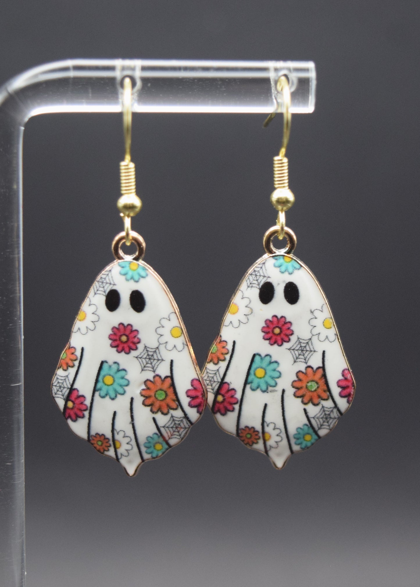 Each earring is shaped like a ghost, with a white base color. The surface of the ghost is decorated with a vibrant pattern of small, colorful flowers in shades of teal, orange, pink, and yellow. Interspersed among the flowers are delicate black spiderwebs, adding a subtle spooky touch to the otherwise cheerful floral design. Each ghost has two prominent black, oval-shaped eyes.