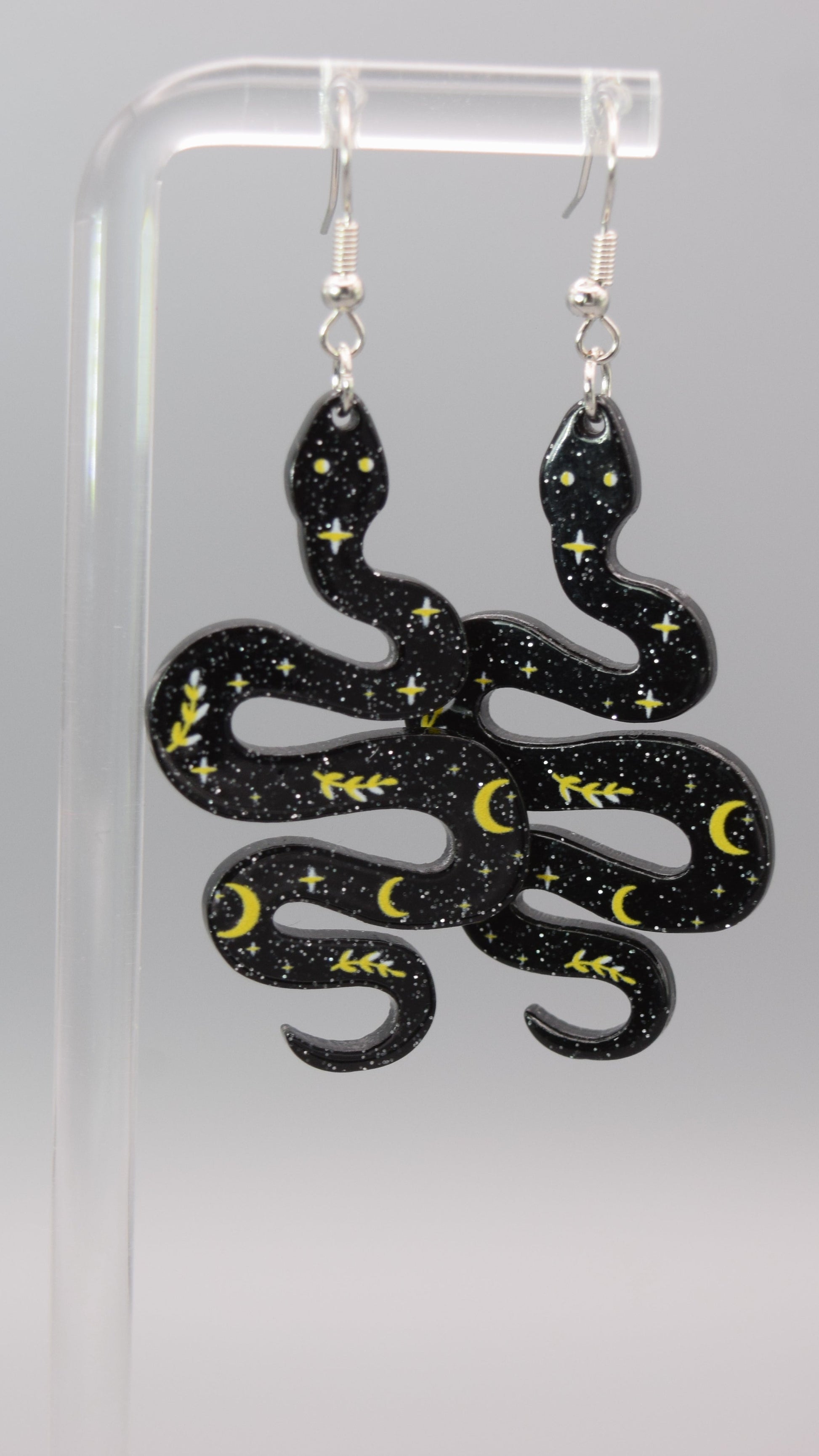 Each earring is shaped like a winding snake, made of a dark black epoxy with silver sparkles in it. The surface of the snakes is adorned with small yellow/ gold numerous tiny stars, crescent moons, and leafy branches. The patterns are scattered across the snake's body, creating a starry night sky effect. The snakes also have two small, light-colored dots for eyes near their heads. The dangle earrings, displayed on a clear acrylic stand against a light gray background.