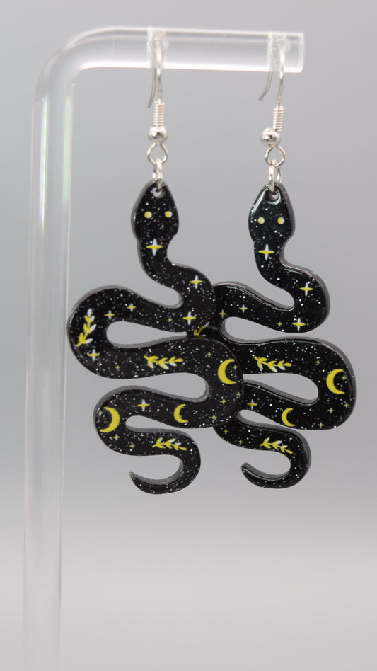 Each earring is shaped like a winding snake, made of a dark black epoxy with silver sparkles in it. The surface of the snakes is adorned with small yellow/ gold numerous tiny stars, crescent moons, and leafy branches. The patterns are scattered across the snake's body, creating a starry night sky effect. The snakes also have two small, light-colored dots for eyes near their heads. The dangle earrings, displayed on a clear acrylic stand against a light gray background.