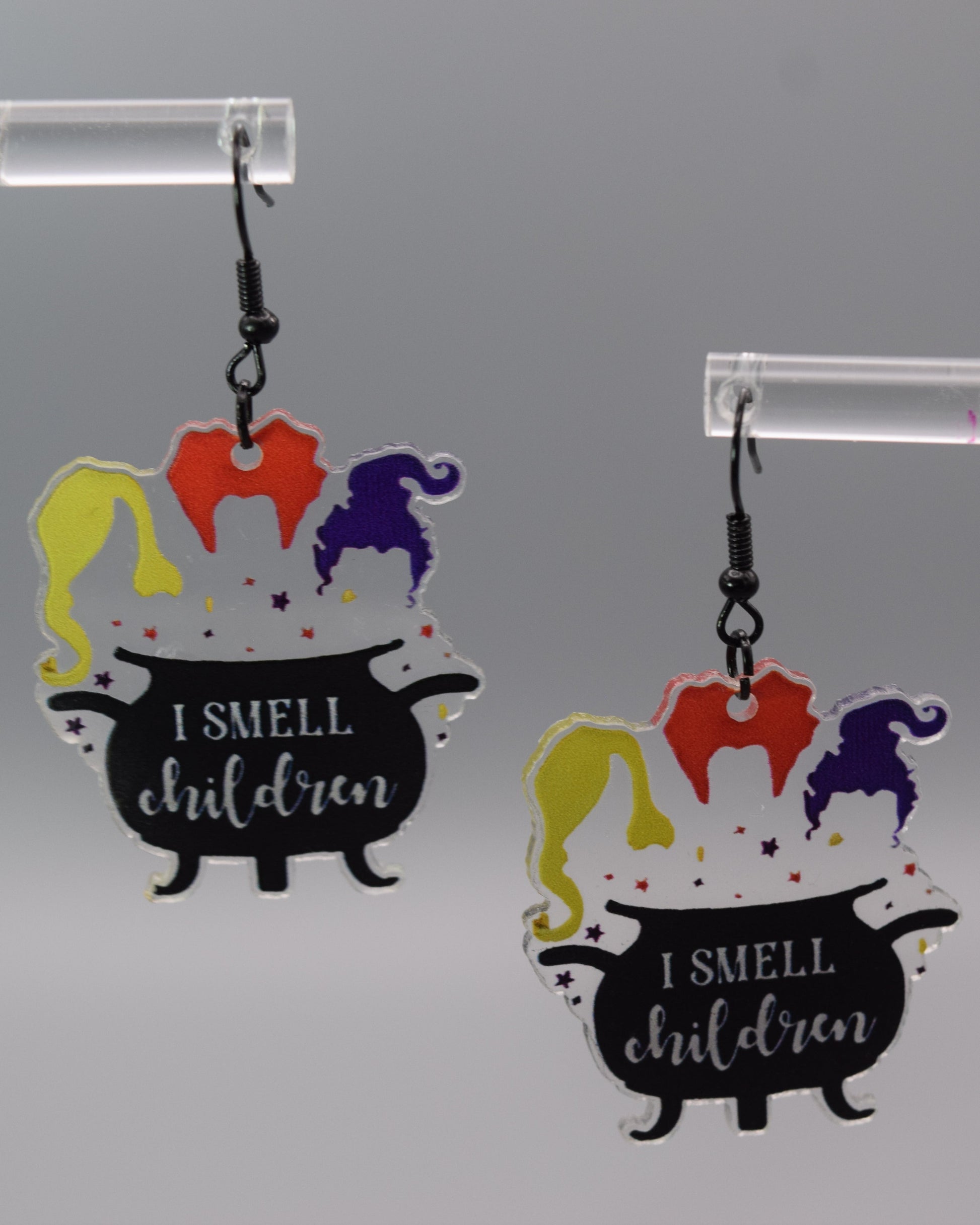 Each earring features a black cauldron with the hair silhouette   representing the three Sanderson sisters: one blonde, one orange, and one purple, with the purple one shaped like a witch's hat. Below the hair, on the body of the black cauldron, white text reads: "I SMELL children". Small purple, yellow and orange stars are scattered around the "steam" and the top rim of the cauldron.