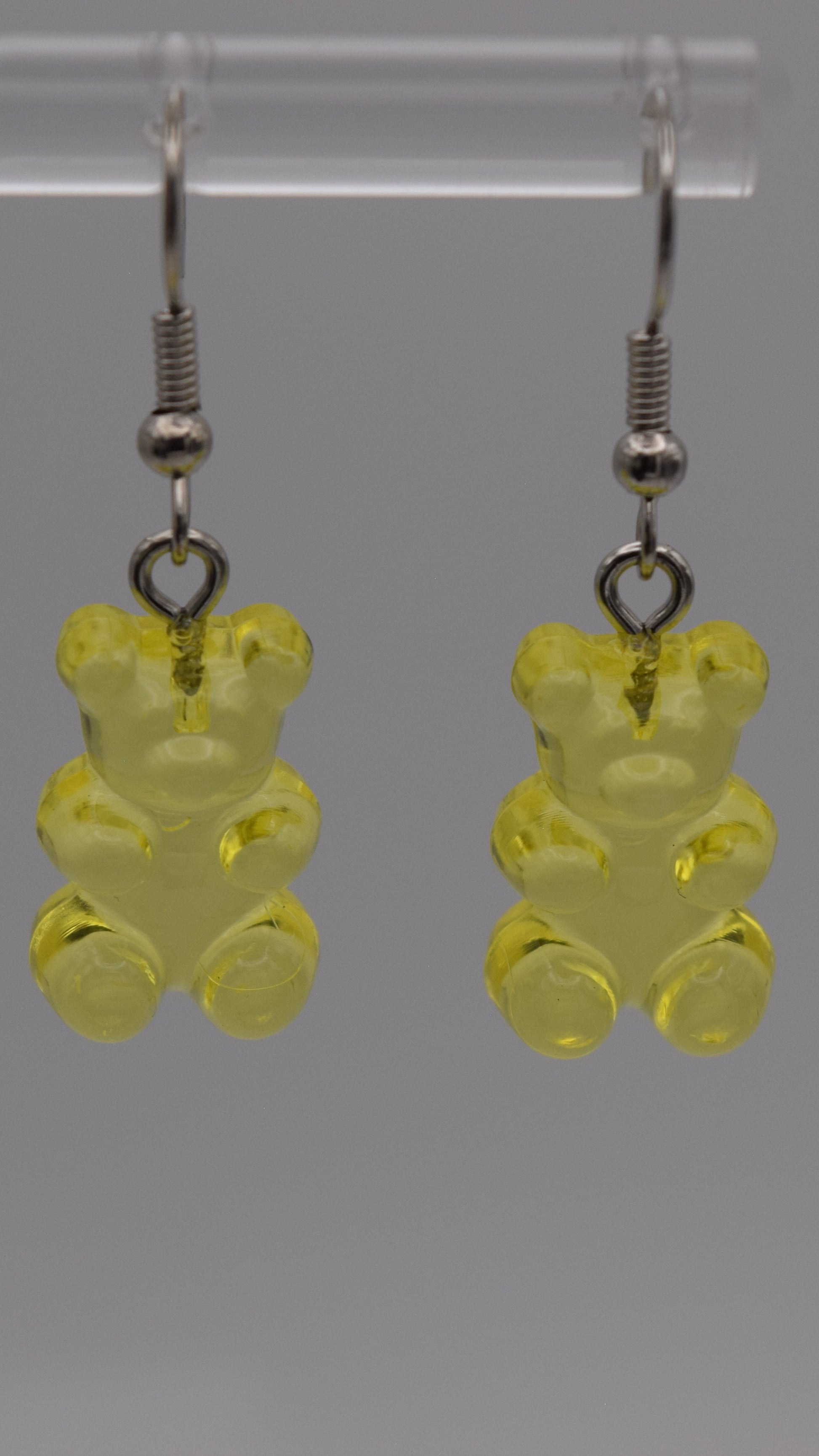 Epoxy 2 inch yellow gummy bear-shaped earrings with silver earring hook. Displayed on a clear stand against a gray background.