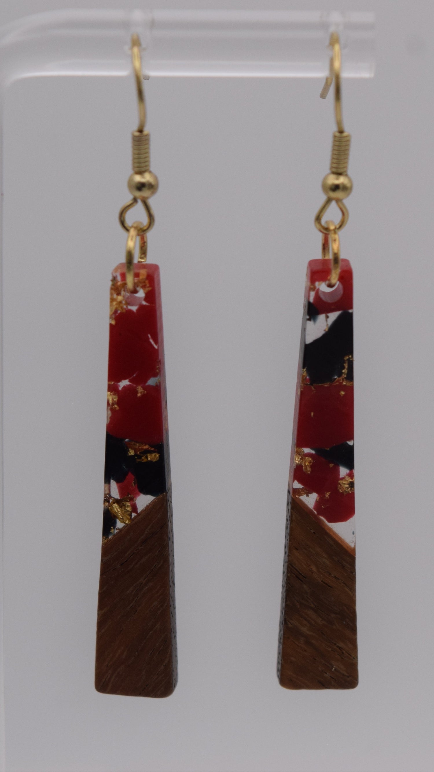 These are a pair of dangle earrings, each earring consists of a long, rectangular drop. The drops are made of two distinct sections. The bottom third of each earring is a solid, dark brown wood, smoothly polished. Above this wooden section, the remaining two-thirds of the earring is made of a clear, glossy resin material. Within this resin, there's a striking abstract design. It's primarily an epoxy mix of clear, red, black, and gold flakes.