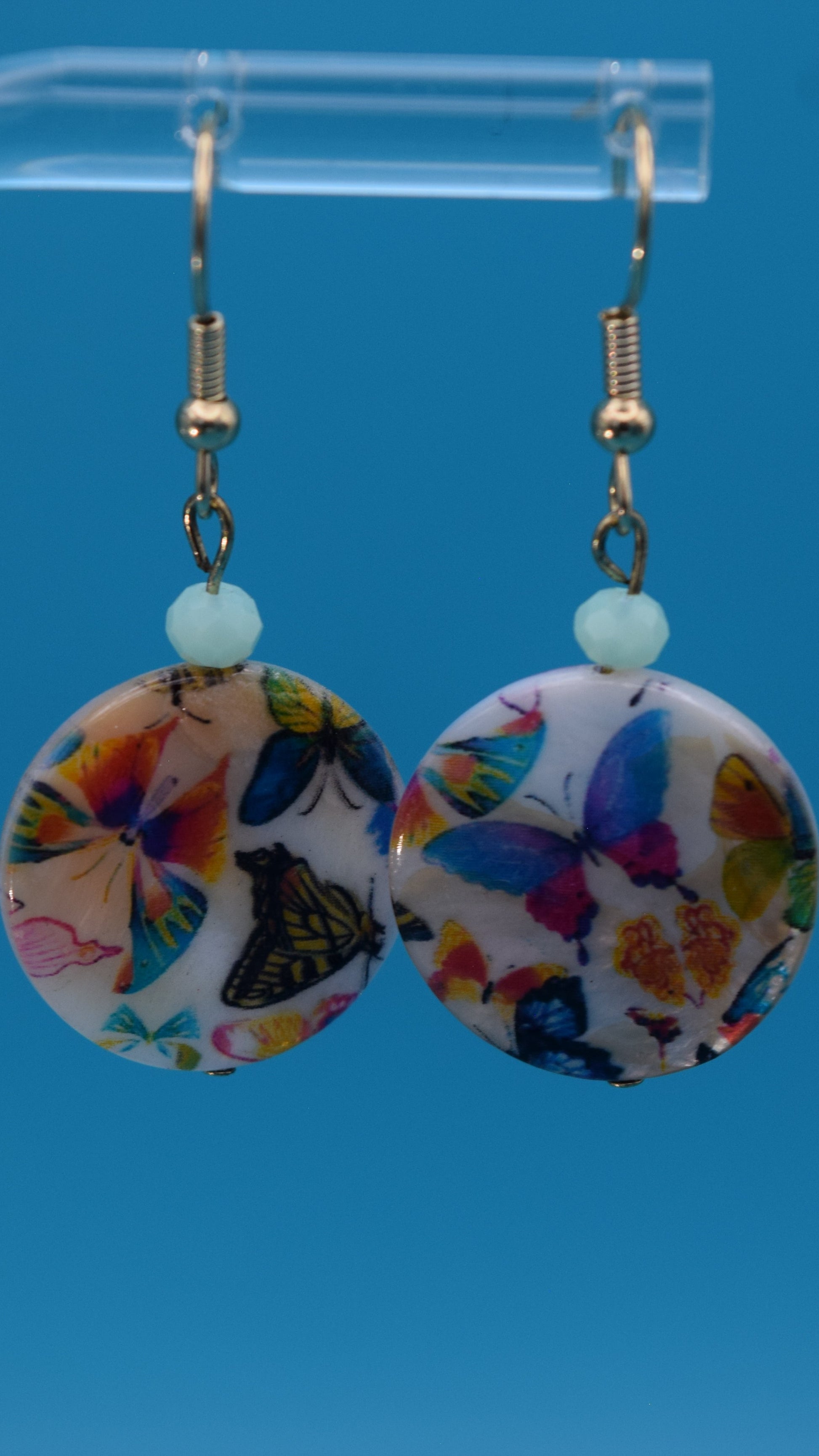 Colorful butterfly-patterned earrings Displayed on a clear stand against a teal background.