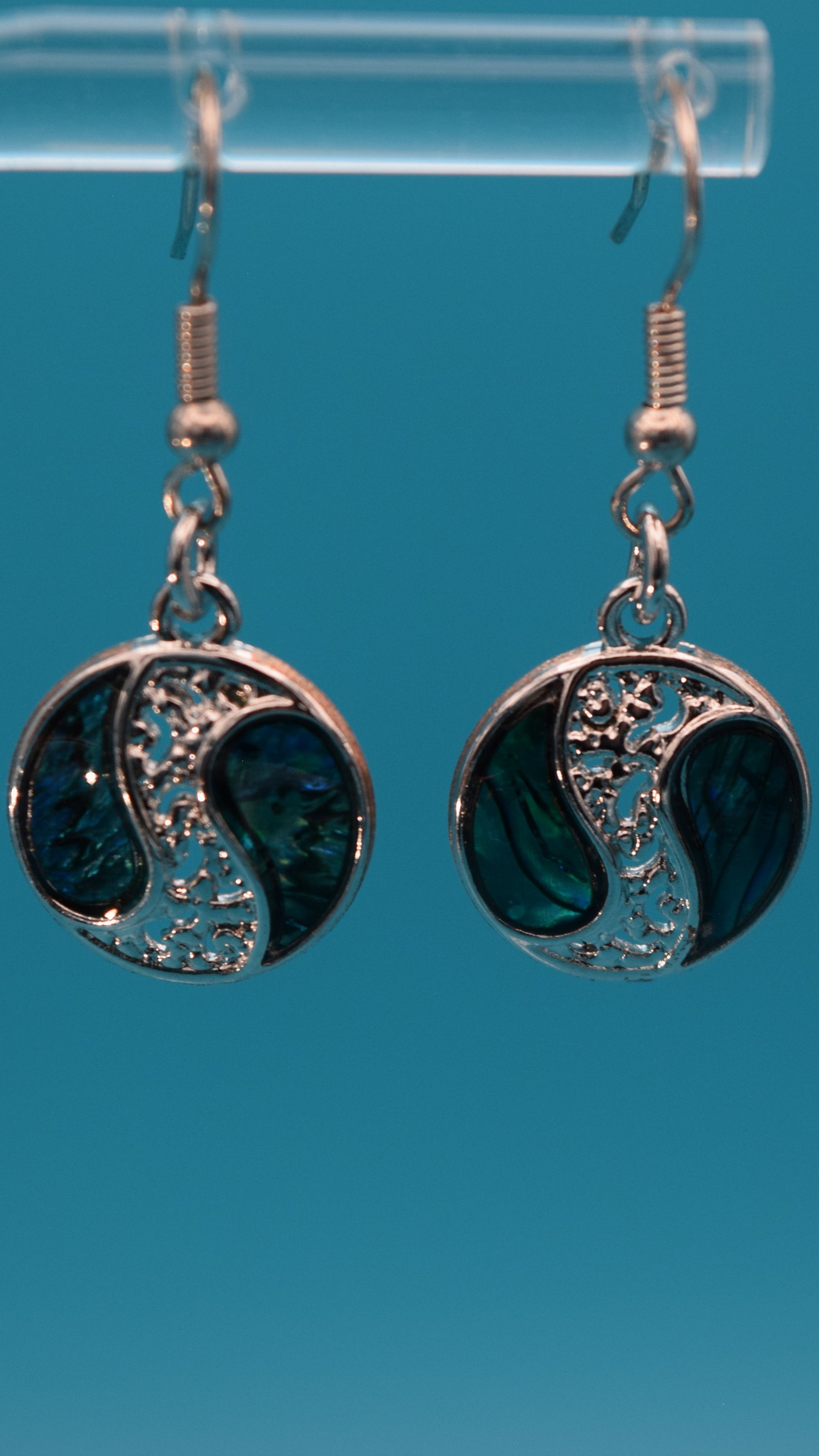 Silver yin yang style earrings dangling 3/4 inch, with iridescent blue, green pattern with black swirls on each side of a silver design. Displayed on a clear stand against a teal background.