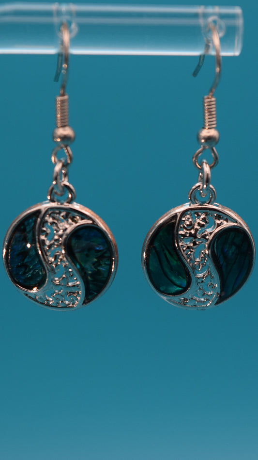 Silver yin yang style earrings dangling 3/4 inch, with iridescent blue, green pattern with black swirls on each side of a silver design. Displayed on a clear stand against a teal background.