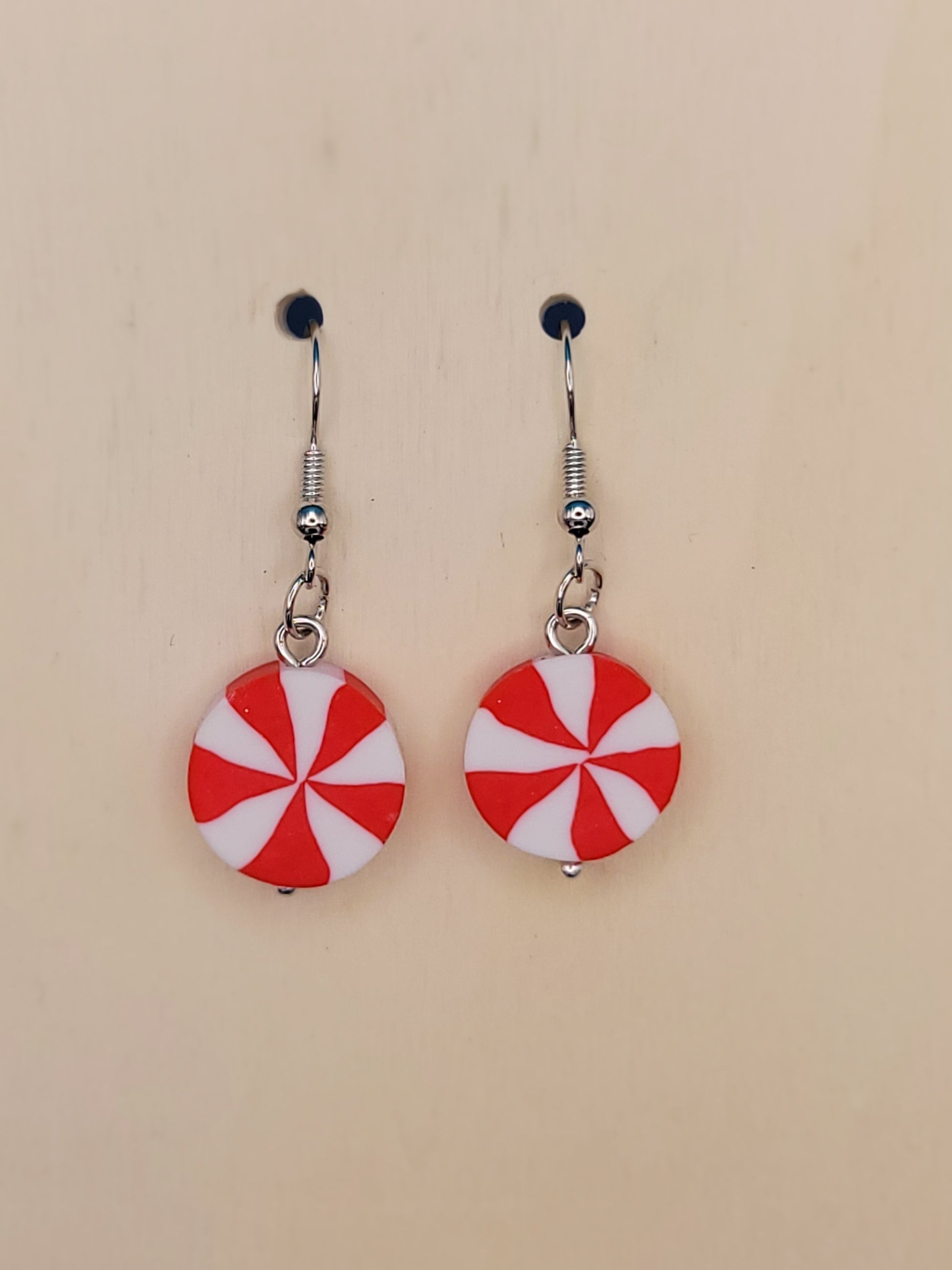 Pair of Silicone red and white peppermint candy dangle earrings. That are 4 cm long with silver hooks and a tiny silver ball on the bottom. Displayed on a beige background.
