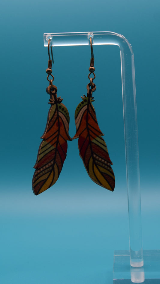 Wooded Feather-shaped earrings colored with a green, orange, read and yellow design, ascended with black lines. Displayed on a clear stand against a teal background